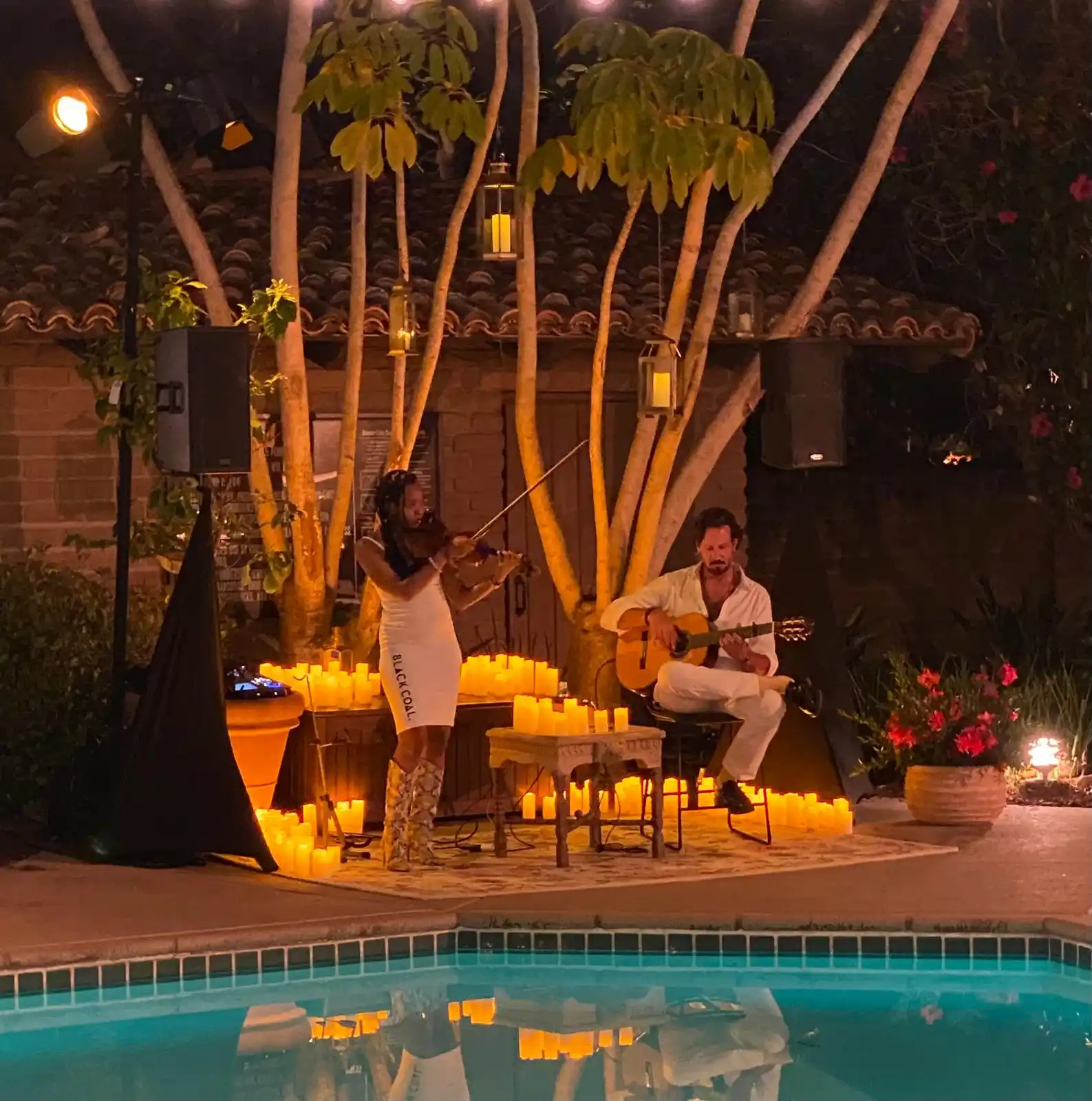 A serene poolside performance with Emilio playing guitar alongside a violinist, surrounded by glowing candles and trees.