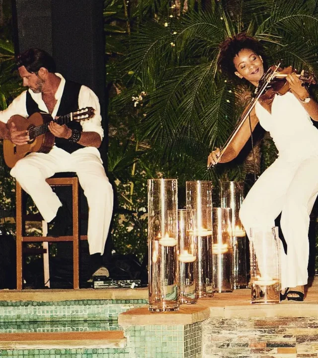 Emilio performing guitar alongside a violinist in a tropical setting, surrounded by elegant candlelight.