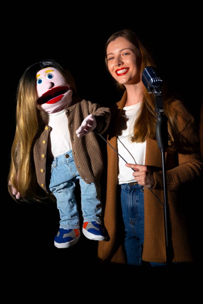 A female ventriloquist holding a puppet during a performance, smiling confidently on stage, capturing the playful energy of a comedy night show.